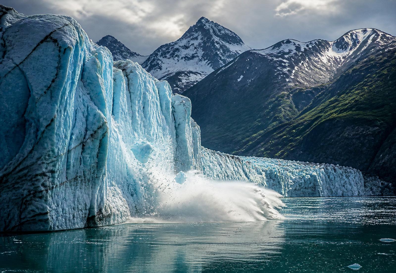 Blue glacier calving into an Alaska fjord with snow-capped peaks